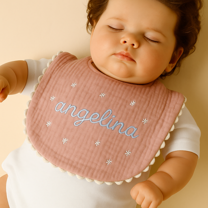 Sleeping baby wearing a pink muslin bib with blue embroidered name “angelina” and white star accents, lying peacefully in a white onesie.