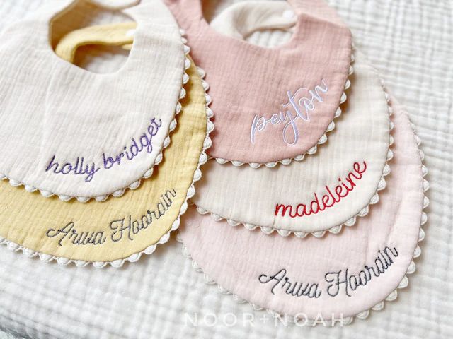 Set of five personalized baby bibs in soft pastel shades with embroidered names and scalloped edges, laid out neatly on a white fabric background.